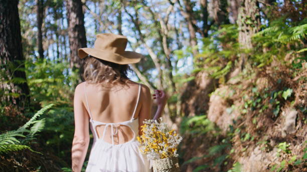 Explorer strolling wild forest overlooking green plants back view. Carefree woman walking jungles wilderness looking on beautiful fern. Female wanderer carrying basket find tranquil beauty in nature
