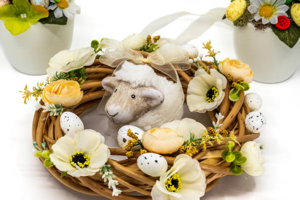 A charming Easter wreath featuring a lamb figurine surrounded by delicate flowers, decorative eggs, and a ribbon bow. The festive arrangement is complemented by additional spring-themed decorations in the background.