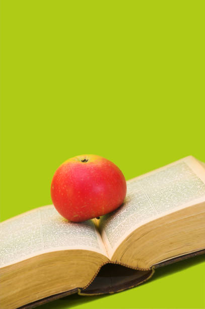 A vibrant photograph showcasing a red apple resting on the pages of an opened book, displayed against a solid bright green backdrop. The image captures a symbolic blend of knowledge and health.