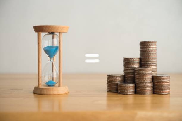 istock Hourglass equal to heap stack coins on wooden table with white wall background. 2197675532