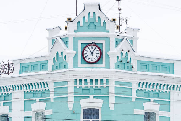A vintage clock tower on a pastel-colored historic building covered in snow, featuring intricate architecture and winter scenery.