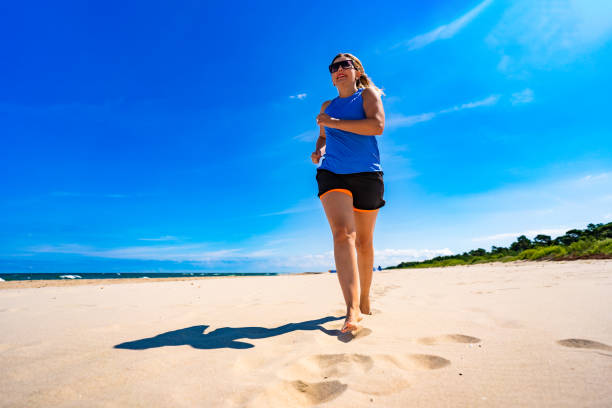 Physical activity on beach in summer. Mature woman running on sandy beach on sunny day. Front view
