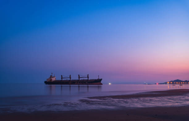 southport, merseyside, united kingdom - 8 september 2020: a container ship passing a solitary figure from the another place installation on the beach reflected in the water at crosby beach in merseyside with a blue sunlit sky