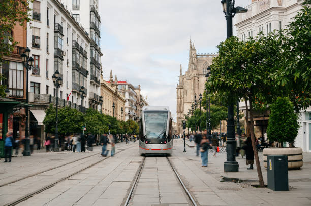 Seville, Spain - March 10, 2025: A modern tram stands on tracks in a historic street while pedestrians walk by. The Giralda Tower can be seen in the background.