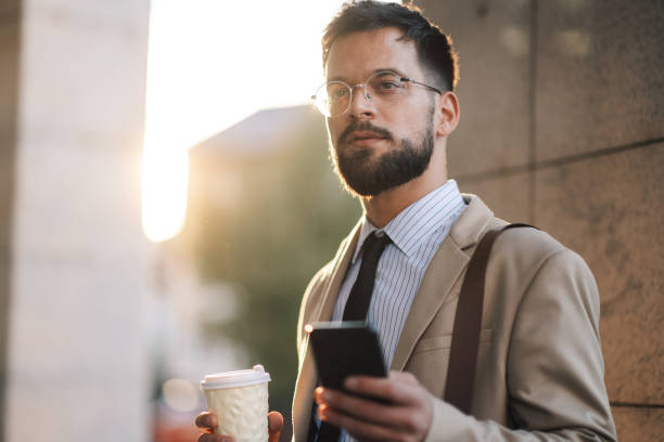 istock Serious bearded businessman using smartphone and holding coffee outside office building 2197738695