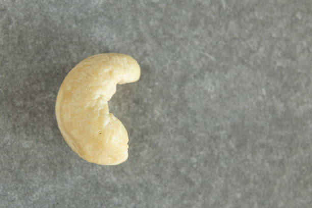 This close-up image features a single cashew nut placed on a grey surface, emphasizing its smooth texture and unique shape. The neutral background highlights the natural, rich details of the nut.