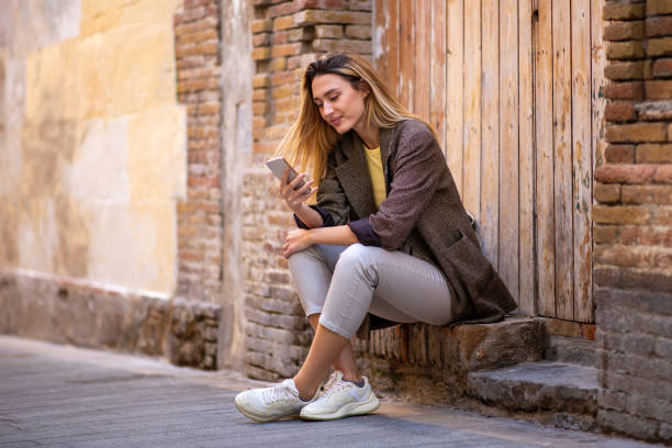 istock Smiling young lady using mobile phone while sitting outdoors against old wooden door 2197743620