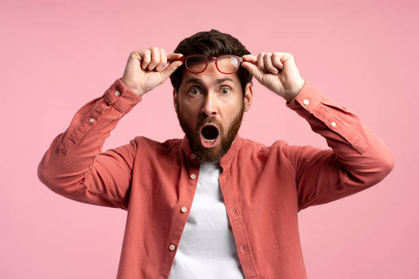 istock Surprised young man taking off in glasses looking at camera while standing against pink background 2197743908
