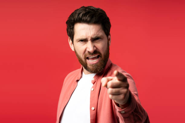 istock Displeased bearded young man looking at camera and pointing by finger, isolated on red background 2197743939