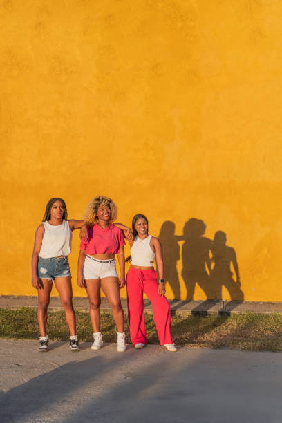 Three female friends are posing together against a vibrant yellow wall, enjoying their time together