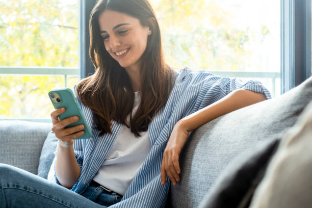 istock Young millennial woman sitting on a couch at home, casually dressed, smiling while using a smartphone to browse the internet, scroll social media, text friends, subscribe to online services 2197747888