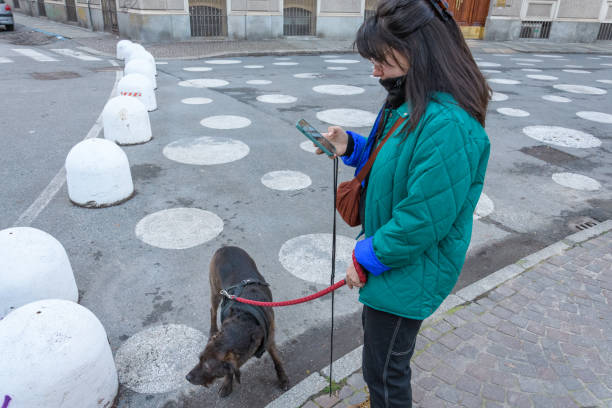 A young Caucasian woman wearing a mint-green jacket that matches her smartphone cover stands on an urban pedestrian zone in Turin. She holds her dog on a leash while intently checking her smartphone. The asphalt is decorated with a playful polka dot pattern, and ample copy space is provided on the left. This image conveys modern lifestyle, digital communication, and pet companionship in a vibrant city setting.