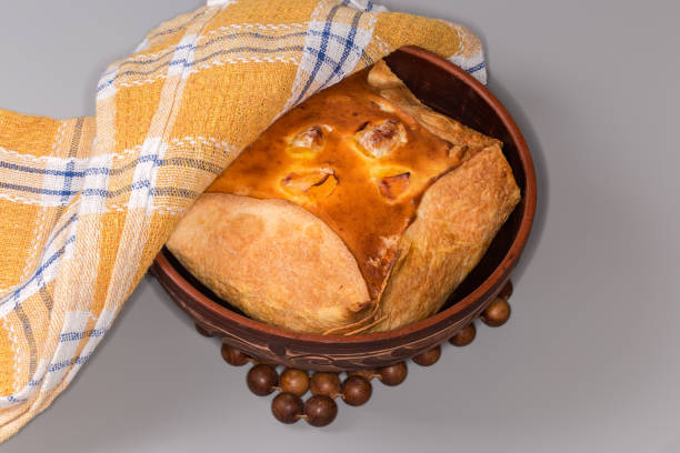 Freshly prepared potato casserole filled with beaten eggs covered with browned crust in flat bread in clay bowl partially under a napkin on a gray background