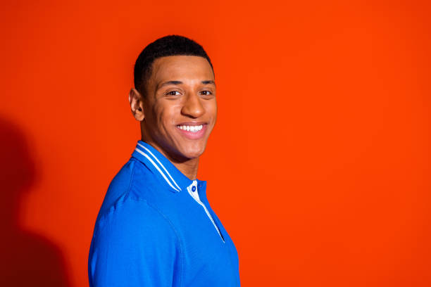 A young, handsome man with black hair in a stylish blue polo shirt smiles happily. Standing against an orange background, he exudes confidence and a trendy lifestyle vibe, perfect for advertising.
