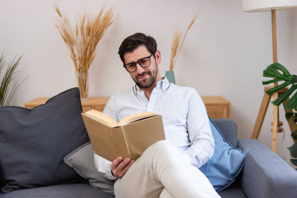 istock Young man reading a book on the sofa at home 2197756260