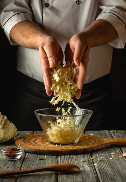 In a warm kitchen, a skilled chef delicately shreds fresh cabbage, letting the pieces fall gracefully into a clear bowl below. The ambiance is filled with culinary creativity and flavor
