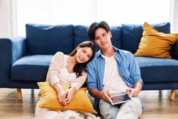istock Young couple relaxes at home, leaning on a cozy blue couch. They smile while using a tablet, enjoying quality time. A warm, stylish living space reflects love, happiness, comfort, and modern lifestyle 2197763085