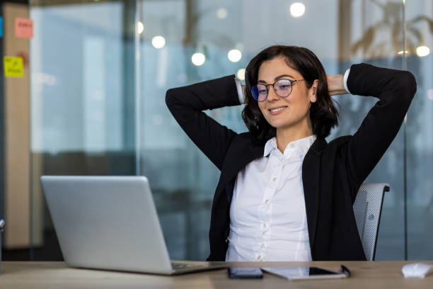 Professional woman in office taking a break while working on laptop, feeling relaxed and confident. Glass office setting provides modern and innovative atmosphere for business creativity