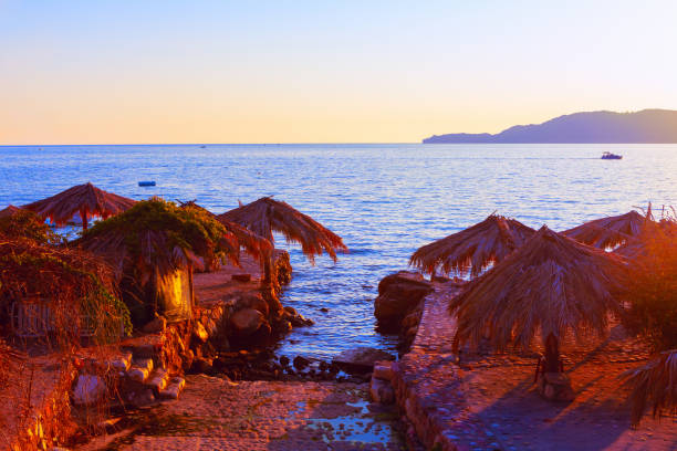 Rustic seaside with straw roofed huts overlooking calm blue waters at sunset. Golden light highlights stone paths and beauty of tranquil coastal retreat, perfect for relaxation and travel inspiration