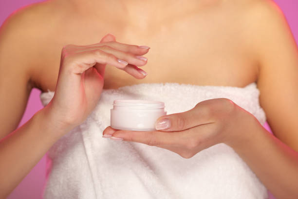 Skincare routine in a bright setting with focus on moisturizer application by a person wrapped in a towel