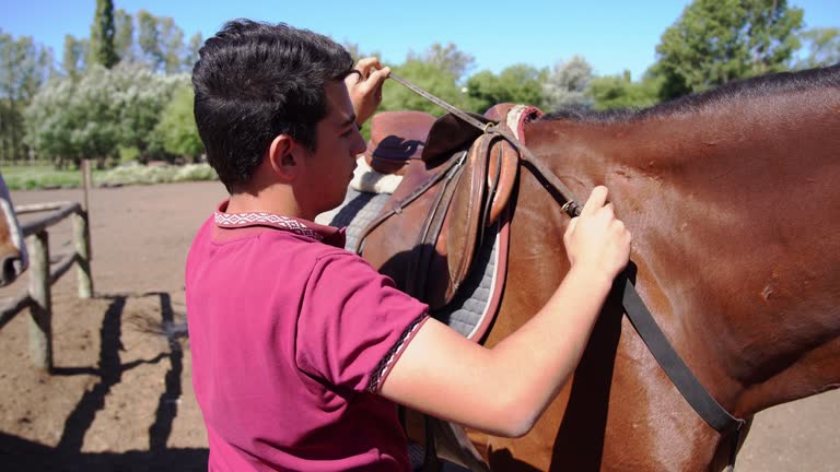 Teenager boy putting the saddle on horse on a ranch