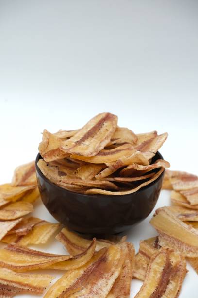 istock Crispy banana chips in a black bowl with scattered pieces around, against white background, showcasing a delicious and healthy snack. 2197788926