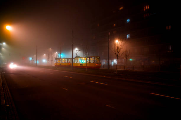 A vintage tram moves through the foggy Warsaw streets at night. The glow of its interior contrasts with the dark surroundings, creating a nostalgic and atmospheric scene in the quiet urban landscape.