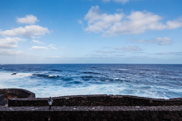 Beautiful ocean view from a volcanic stone wall. The deep blue sea meets a bright sky with fluffy clouds, creating a serene and picturesque coastal landscape.