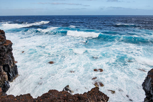 Dramatic ocean waves crashing against the rugged volcanic coastline of El Hierro, Spain. The deep blue sea contrasts with foamy white water, evoking power and nature's force.