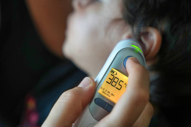 A caregiver holds a digital ear thermometer displaying a temperature of 38.5°C while measuring a child’s fever. The illuminated screen and green indicator light highlight the reading.