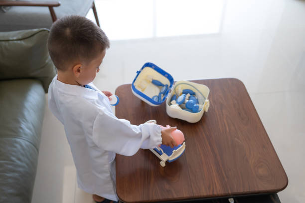 3 year old young boy dressed in a white lab coat pretends to be a doctor, using various toy medical tools on a table in a modern living room, fostering creativity, imagination, and understanding of healthcare roles.