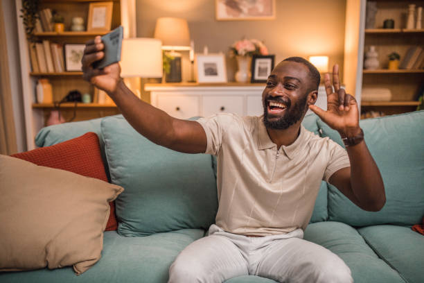 istock Smiling black man taking selfie photo while relaxing in living room 2197807092