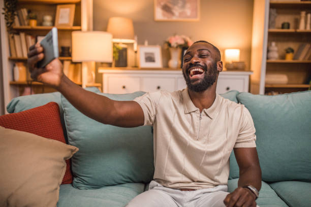istock Smiling black man taking selfie photo while sitting on sofa in living room 2197807127