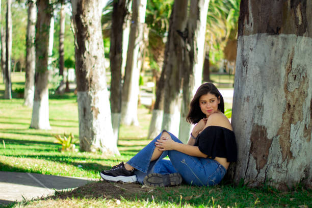 Woman sitting on the ground with green grass and leaning on a tree trunk. Calm Green Space