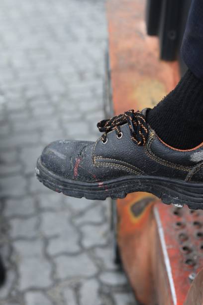 Detailed close-up of a worn black work boot with orange laces, showing signs of use and wear, resting on an industrial surface with a textured background. Ideal for themes of labor and industry.