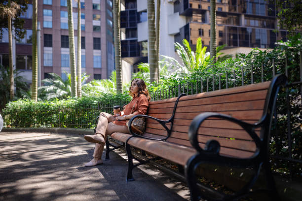 A woman enjoys a peaceful moment on a park bench in Sydney, surrounded by lush tropical plants. She sips a warm beverage, fully immersed in the serene atmosphere.