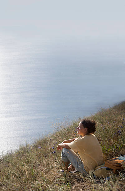 An Asian woman tourist stands at a viewpoint, taking in the breathtaking seascape view of Bali, Indonesia, with a clear blue sky above.
