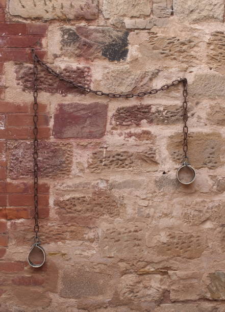Rusty iron shackles hanging on an aged stone wall, evoking historical imprisonment and medieval architecture. Weathered textures and muted tones add a haunting, rustic atmosphere