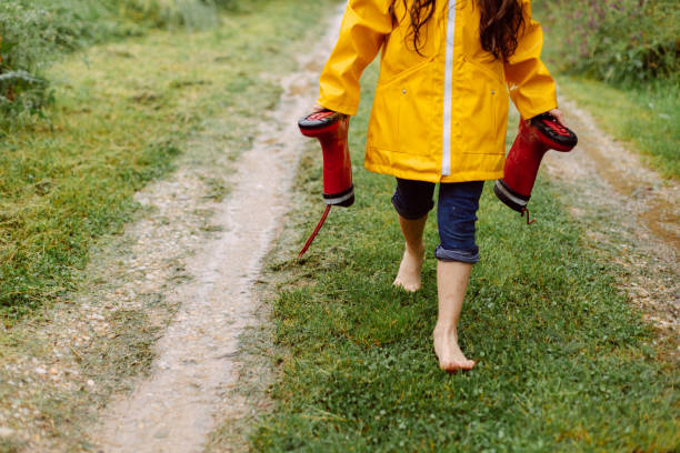Joyful young girl standing barefoot in a muddy puddle on a rural dirt path during a light rain. She is wearing a bright yellow raincoat. Her long, wet hair drapes over her shoulders while she is enjoying the moment. She holds a pair of red rain boots in her hands ,adding a playful touch to the scene.