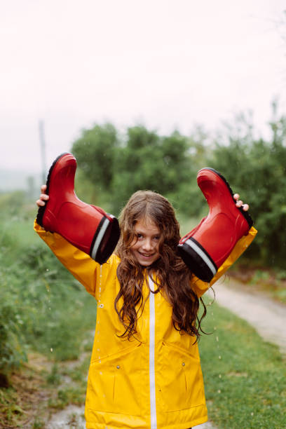 A young girl joyfully playing in a muddy puddle on a rural dirt path during a light rain. Dressed in a bright yellow raincoat, she holds up her red rain boots, pouring out water from them with a playful expression. The lush greenery surrounding the path enhances the carefree and adventurous spirit of childhood fun in the rain.