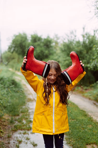 A young girl joyfully playing in a muddy puddle on a rural dirt path during a light rain. Dressed in a bright yellow raincoat, she holds up her red rain boots, pouring out water from them with a playful expression. The lush greenery surrounding the path enhances the carefree and adventurous spirit of childhood fun in the rain.