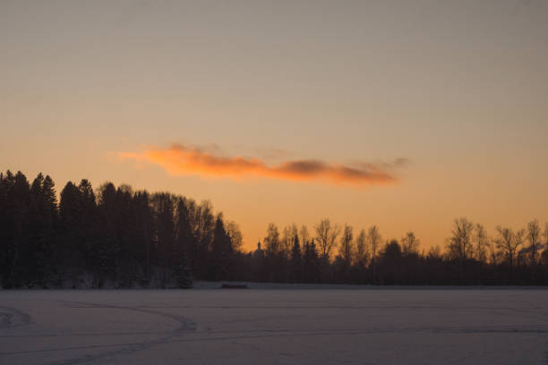 Beautiful winter landscape with surface of the frozen lake in the foreground and silhouettes of trees on the orange evening sky background. The silhouette of old church is visible on the horizon