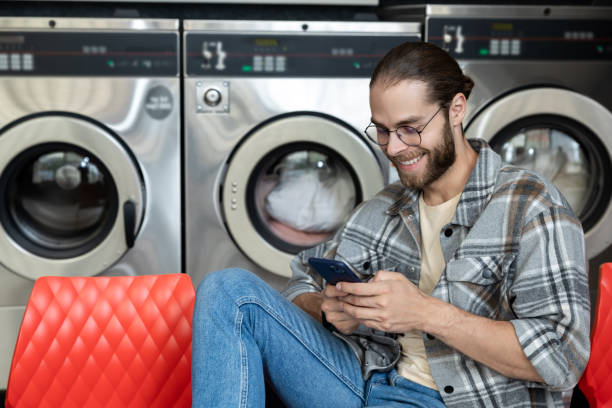 istock Bearded man in laundromat using washing machine while browsing online on mobile phone 2197861380