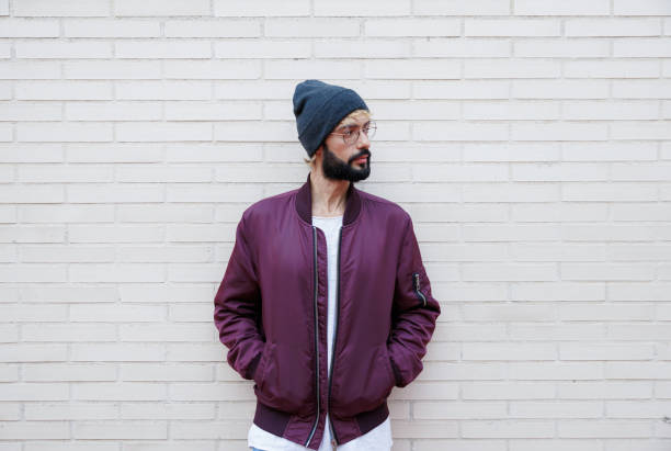 Portrait of a trendy man wearing a purple bomber jacket and beanie, standing against a white brick wall with hands in his pockets