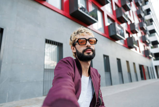 Trendy influencer sporting dyed hair and a beard is capturing a selfie in front of a sleek, modern building in an urban setting