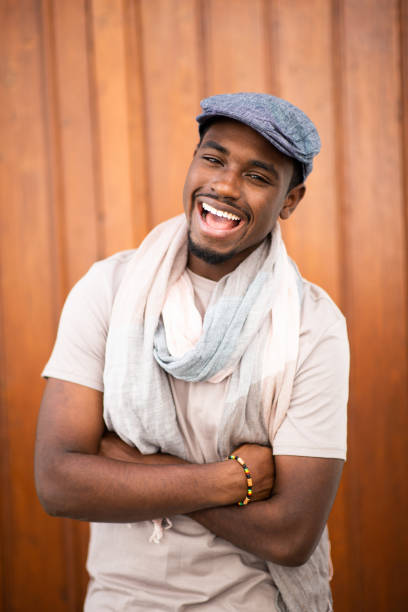 Portrait of laughing young african american man with cap and scarf against wooden background with arms crossed