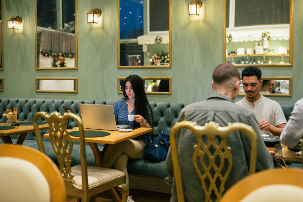 Wide angle shot of young Middle Eastern woman working remotely and drinking coffee sitting at a fancy cafe by a group of multicultural young people