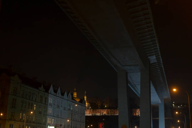 A person is standing quietly under an old bridge at night, observing their surroundings while the darkness envelops the city around them