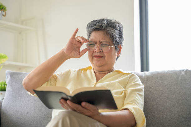 An elderly woman has an eye long sighted problem sitting on a couch adjusts her glasses while reading a book. Senior lifestyle, vision care elder people lifestyle health care problem concept.