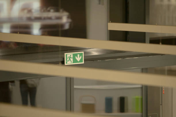 A green emergency exit sign featuring a running figure and an arrow pointing right, mounted on a red ceiling with skylights. The sign is illuminated, indicating its readiness for use. The visual style is modern and functional, conveying urgency and safety. Potential usage includes architectural design references, safety compliance documentation, or educational materials about emergency procedures.